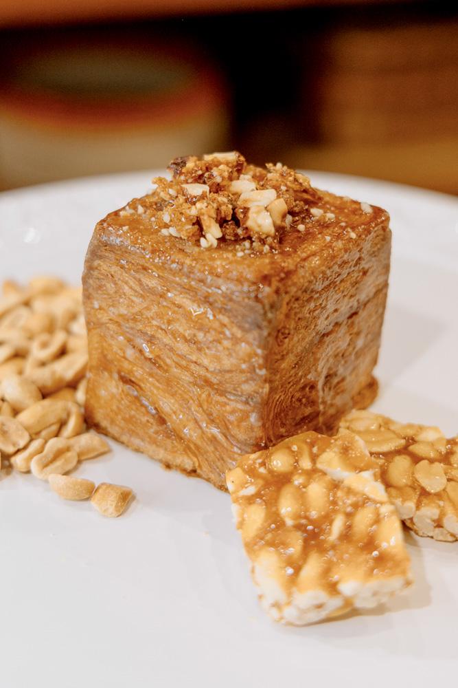 Doce em forma de cubo, coberto por pedaços de amendoim e rodeado por pedaços de pé-de-moleque sobre um prato raso de cor branca