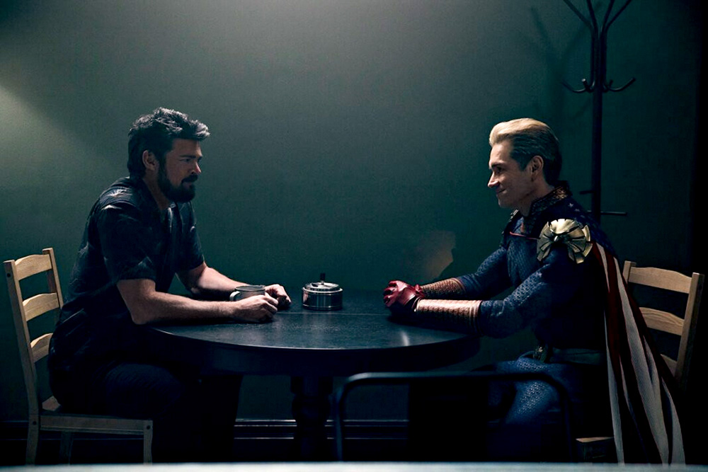 Imagem mostra dois homens sentados em uma mesa, em uma sala escura. O da direita veste uma roupa de super-herói.