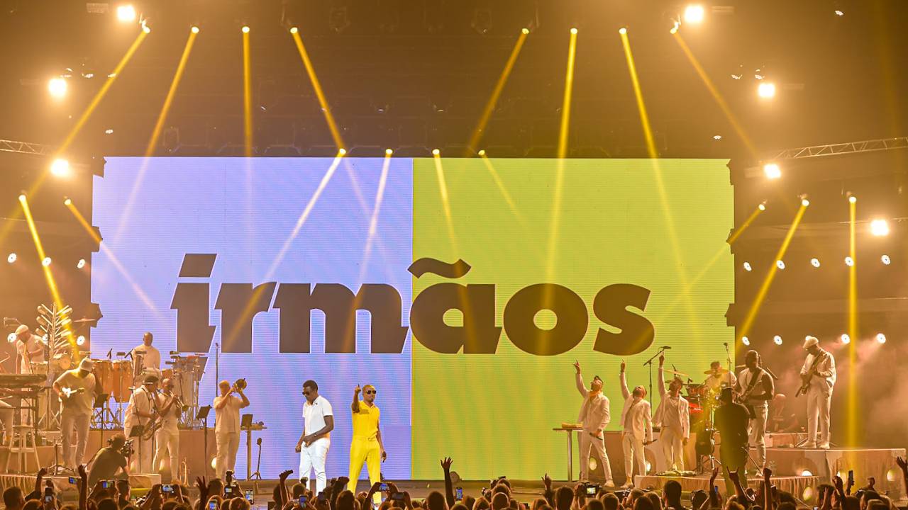 Imagem mostra dois homens, um de roupa branca e o outro de roupa amarela, no meio de palco em frente à multidão e entre músicos.