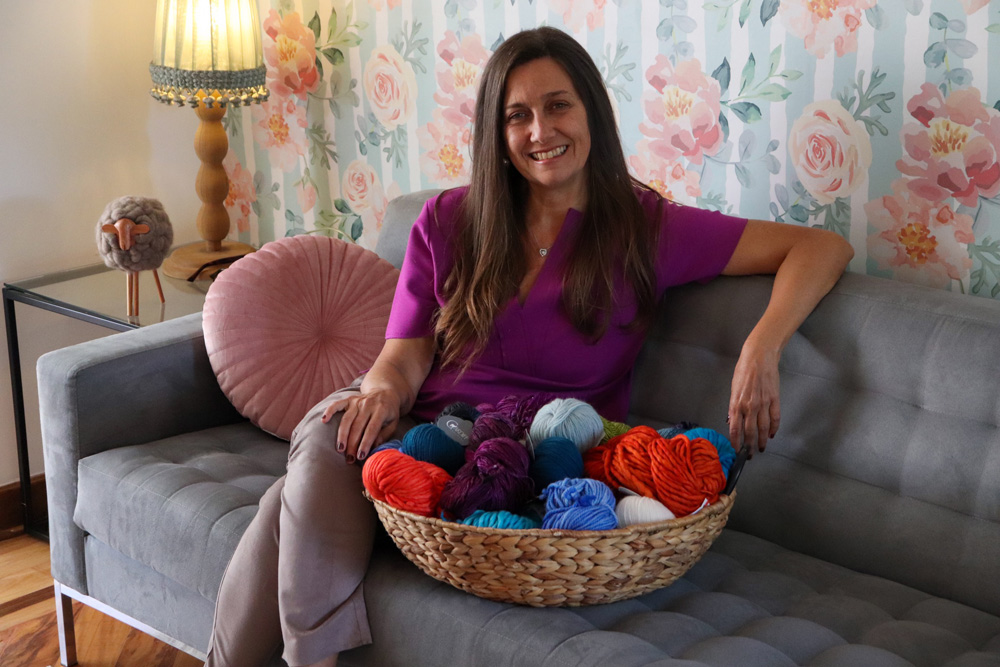 Aida Fonseca posa sentada em sofá cinza com cesta repleta de novelos de lã. Ao fundo, abajur está ligado e o papel de parede tem rosas.