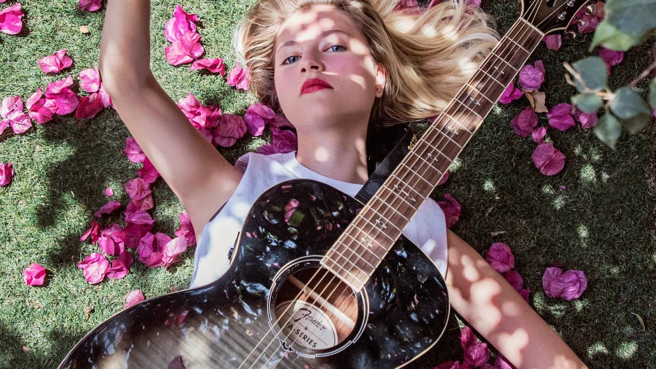 Ana Schurmann posa deitada com violão preto em cima do corpo e rosas espalhadas pelo gramado. Está com uma mão erguida e os cabelos loiros no chão.