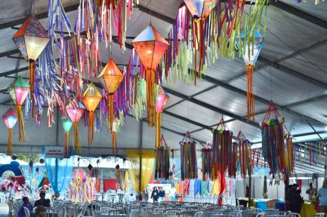 Balões de papel pendurados no teto de um grande salão, onde são vistas também mesas e cadeiras de plástico