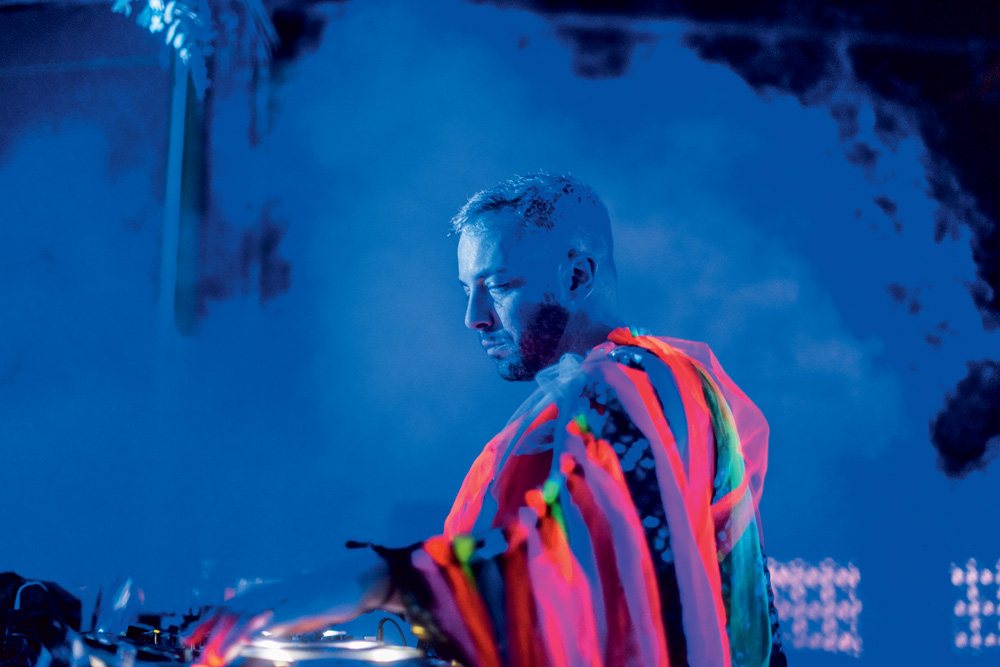 Augusto Olivani, fundador do selo Selvagem, aparece discotecando de lado, com roupa de listras coloridas e luz azul na parte de cima da imagem.