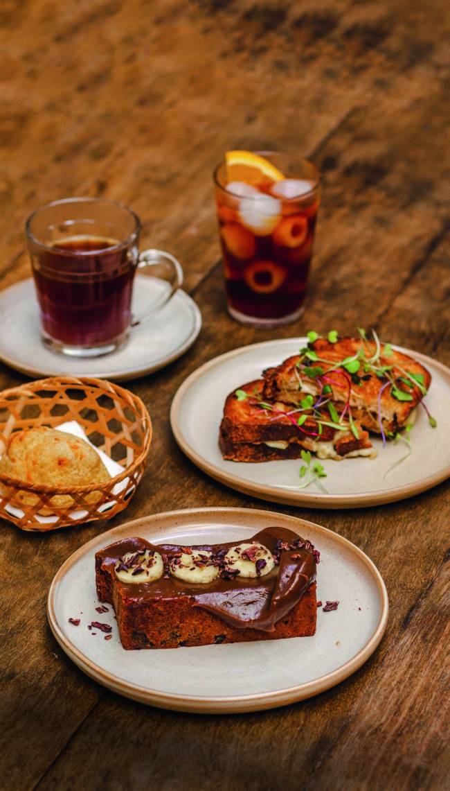 Queijo quente, bolo de banana, pão de queijo e cafés dispostos sobre uma mesa de madeira