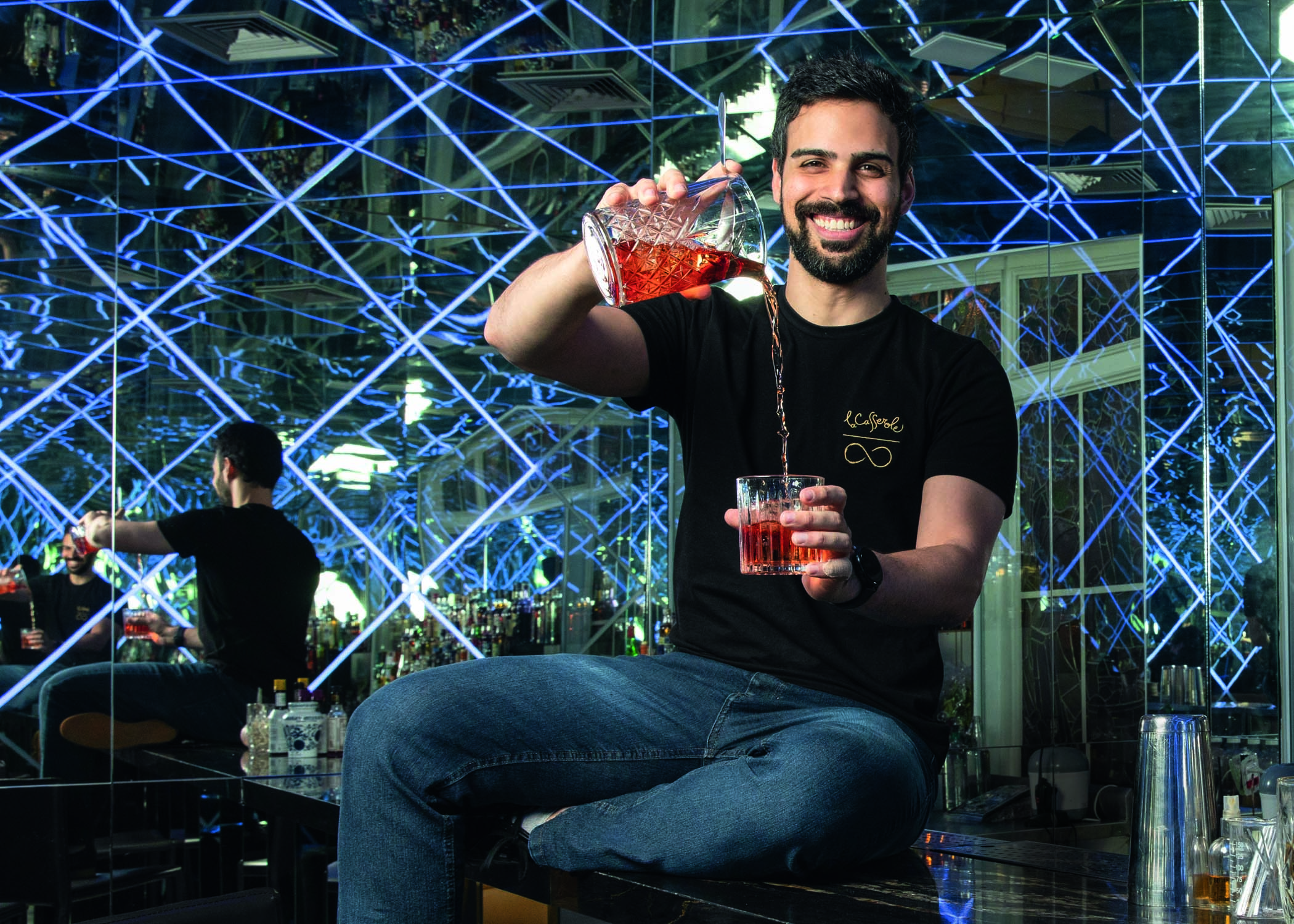 Henry, dono de dois bares, sentado colocando um drinque vermelho de uma jarra de vidro em um copo de vidro. Ele é branco, magro, de cabelos e barba escuros. Sorri para a foto e está sentado dentro de um de seus bares, repleto de luzes azuis