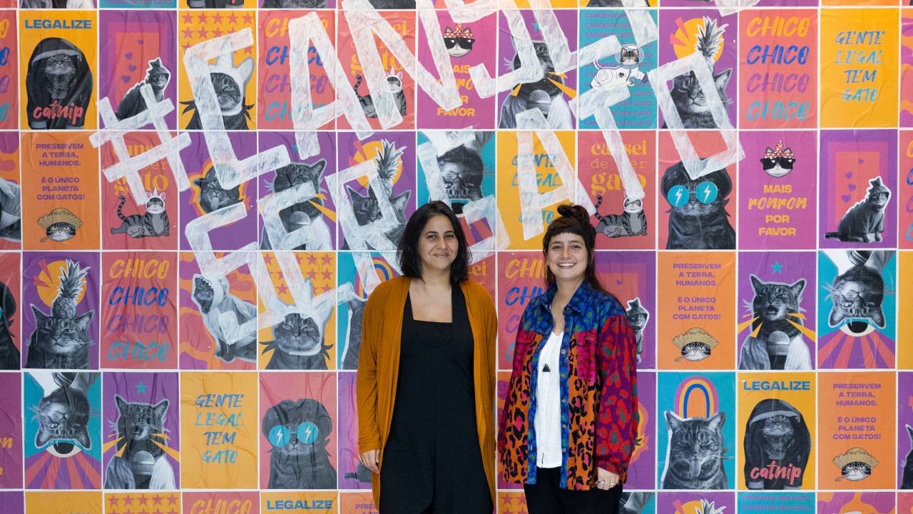 Em frente a grande parede com lambes coloridos, duas mulheres posam de pé lado a lado.