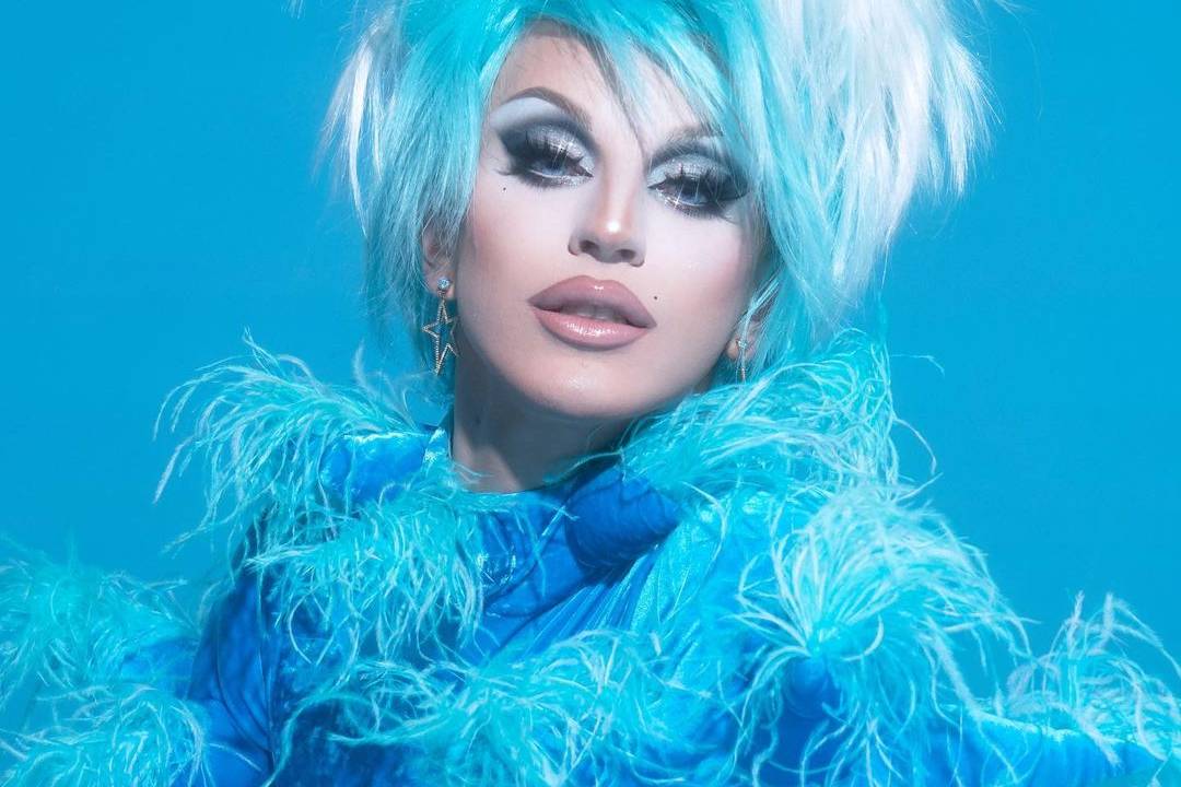 A drag queen Aquaria aparece toda de azul com plumas e cabelo alto. Está com maquiagem bem branca e batom nude.