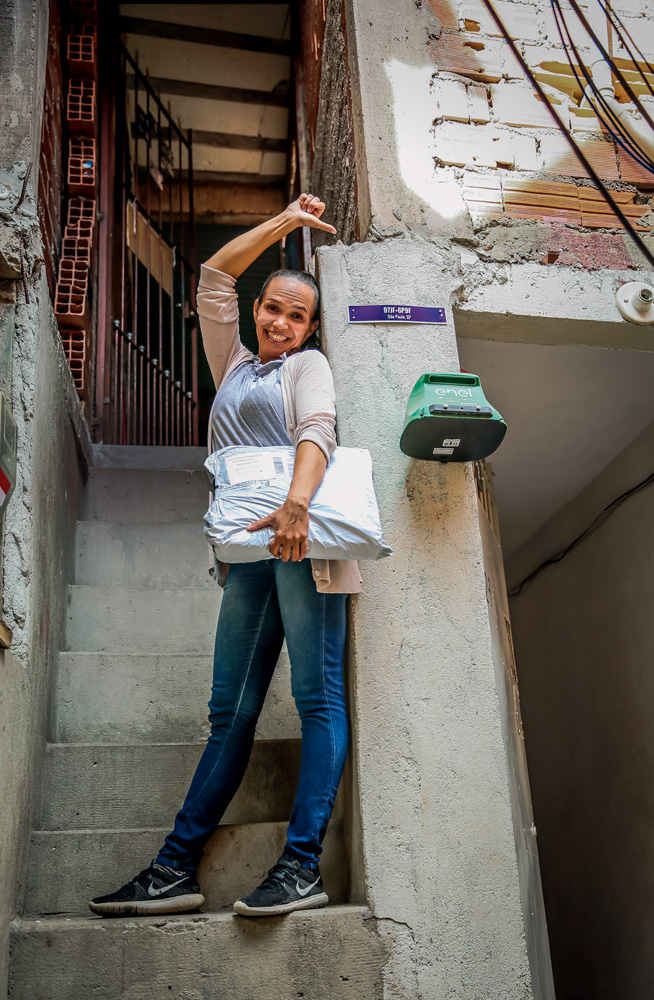 Alexandra, mulher negra e magra, na frente de sua casa, em Paraisópolis. Segura um pacote de encomenda on-line e aponta para a placa de seu endereço