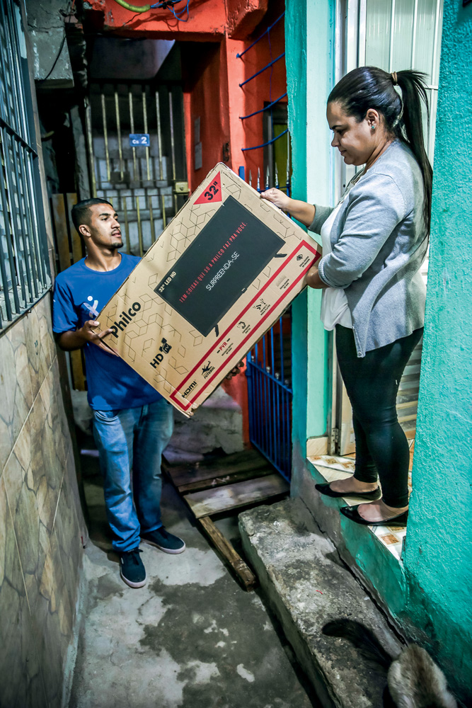 Um entregador, com camiseta azul, seu uniforme, entrega uma grande caixa de papelão a um mulher na porta de uma casa, em Paraisópolis