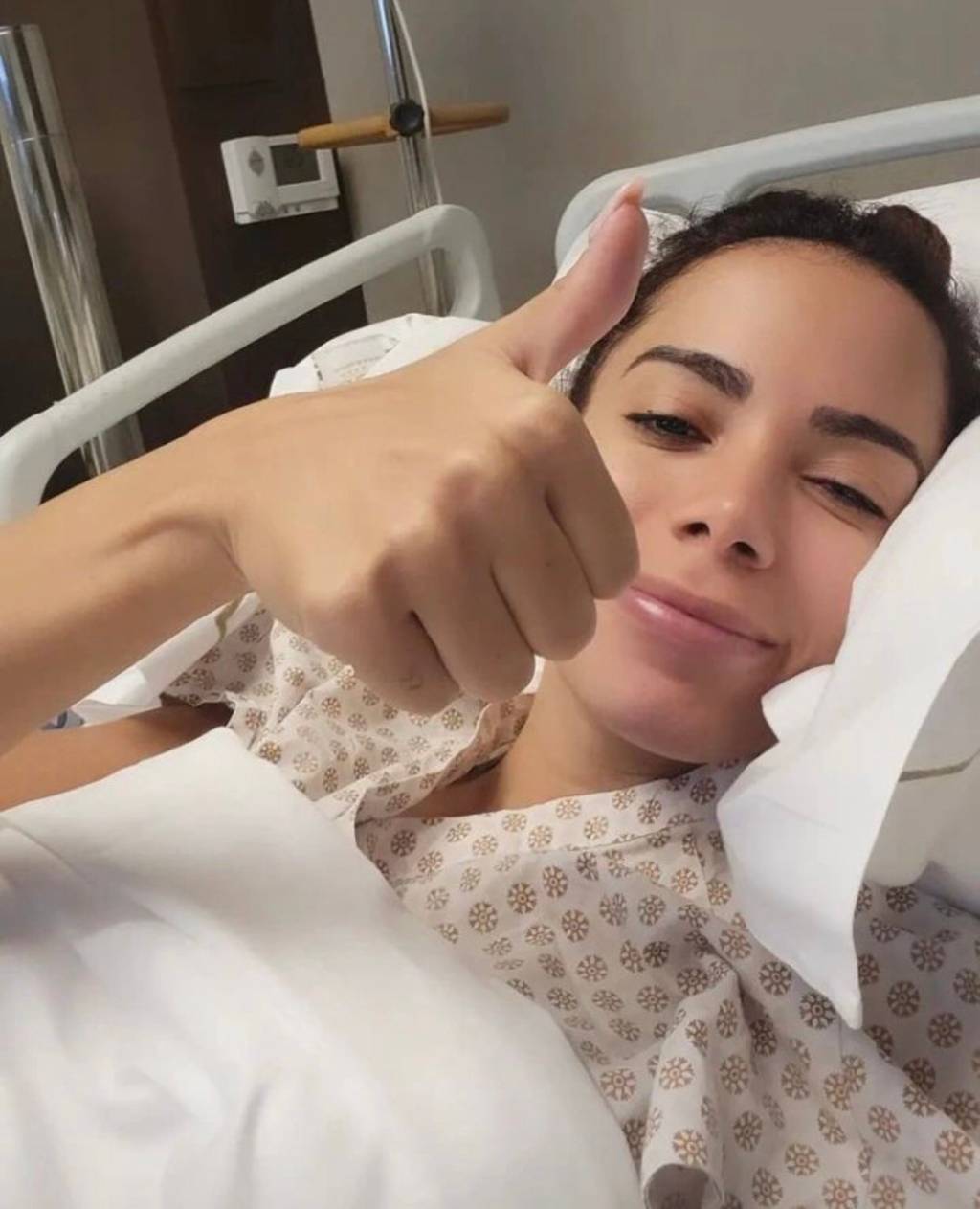 A cantora Anitta, deitada em uma cama de hospital, faz um símbolo de joia com o dedo da mão.