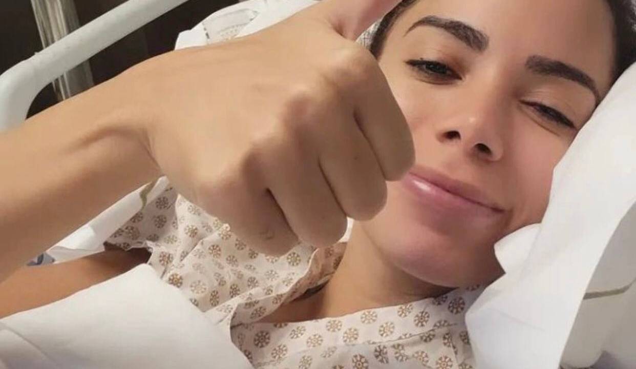 A cantora Anitta, deitada em uma cama de hospital, faz um símbolo de joia com o dedo da mão.