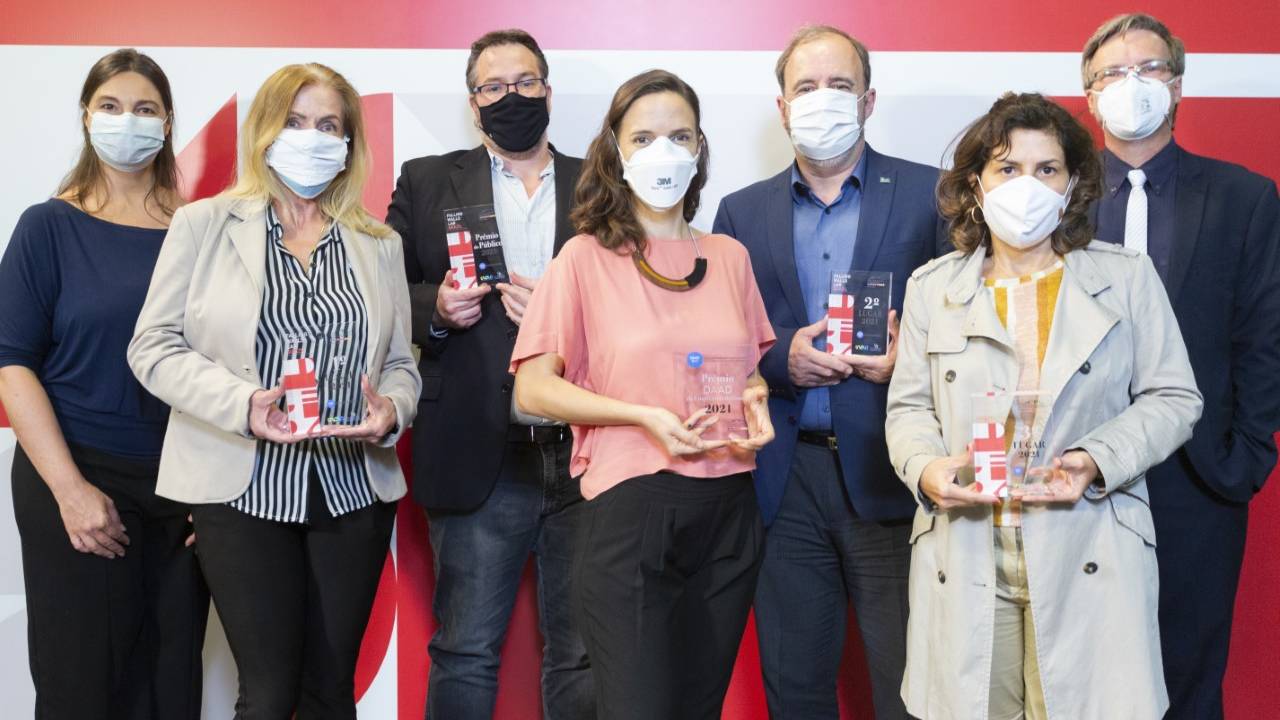Cinco pessoas posam de pé, lado a lado, com cartaz branco e vermelho ao fundo. Todas usam máscara.