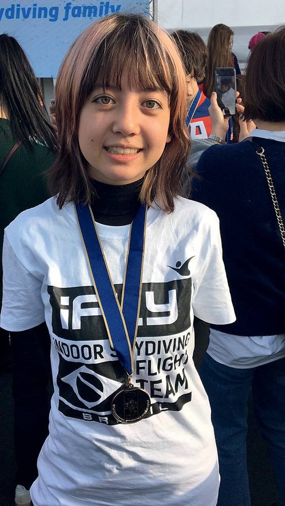 Jovem sorri para a foto com medalha no pescoço e camiseta branca.