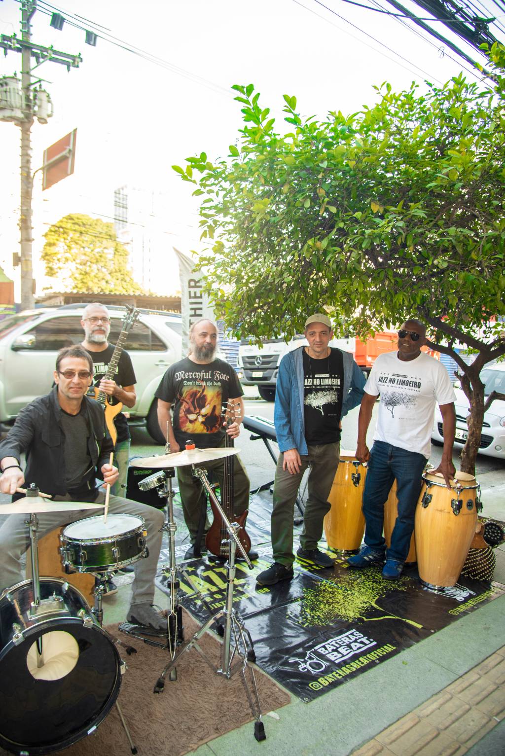 Cinco homens posam debaixo de pé de limão com instrumentos e carros ao fundo.