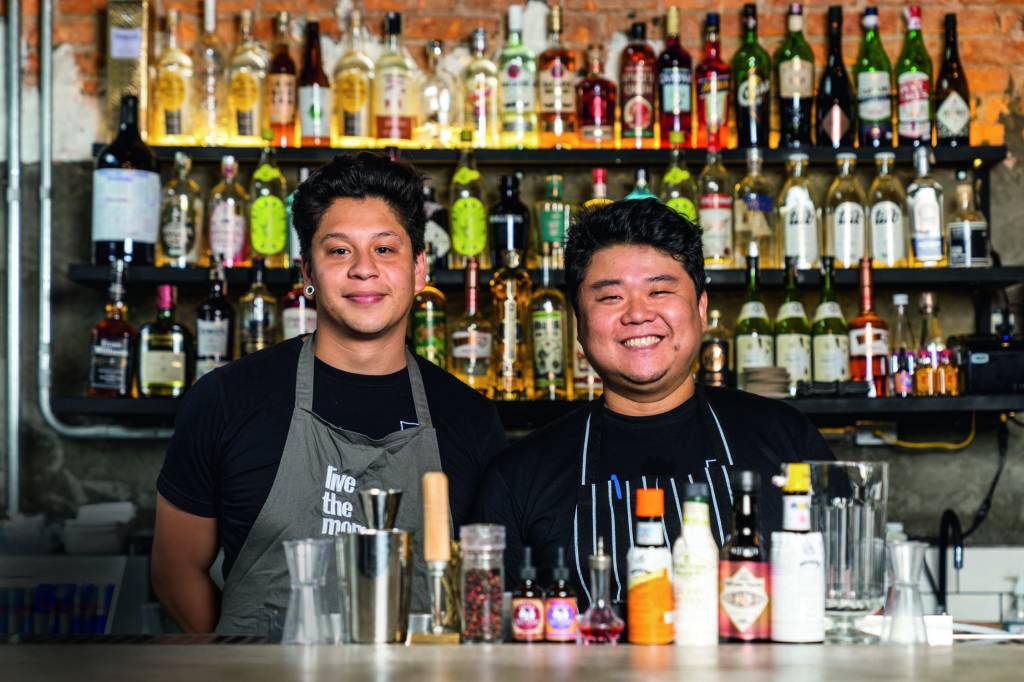 Dois homens, um branco e outro com traços asiáticos, atrás de um balcão de um bar, com várias garrafas ao fundo
