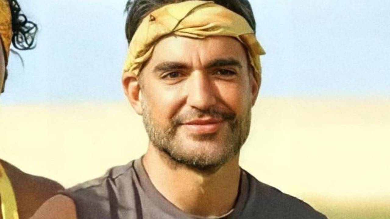 Matheus aparece com bandana amarela na cabeça, regata preta e cabelos pretos no alto. Sorri sem mostrar os dentes e tem barba por fazer.