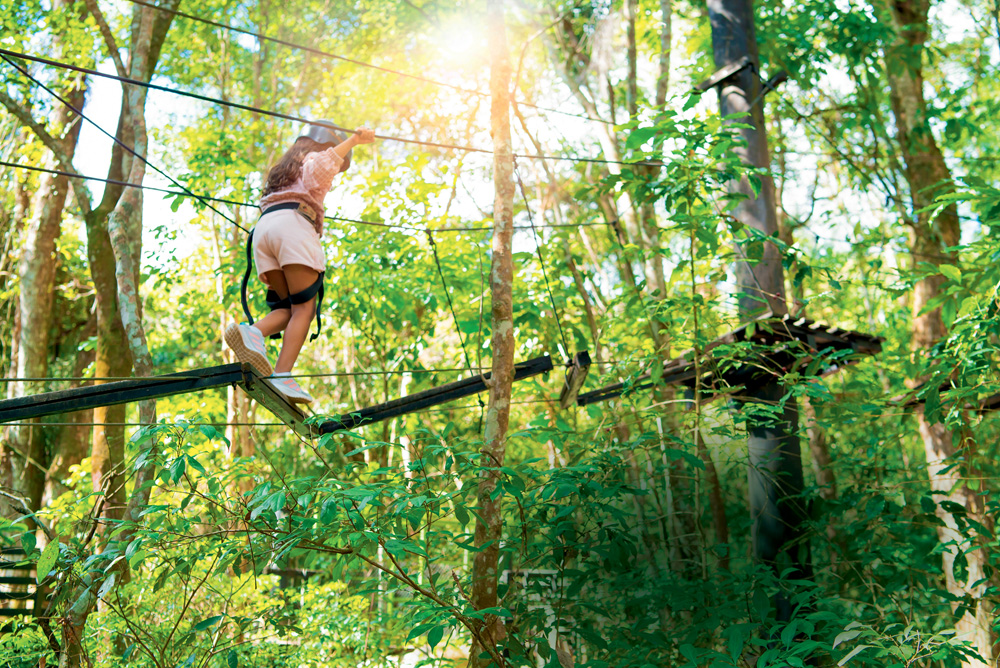 Menina brinca em ponte suspensa nas árvores. Ela usa equipamentos de proteção