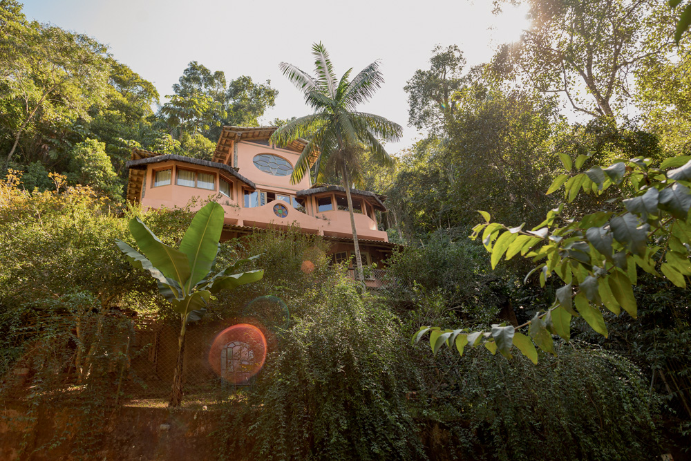 Fotografada de baixo, casa com tons terrosos aparece em meio a árvores e palmeiras.