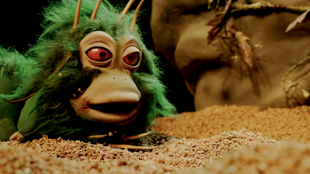 Boneco que parece um lagarta, com olhos vermelhos e pelo verde. Ele está em um palco com areia