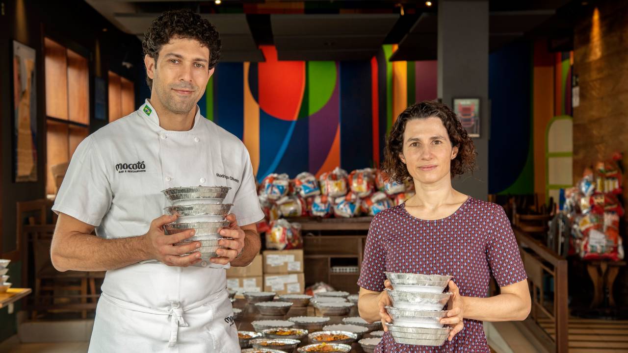 O chef Rodrigo Oliveira segurando quentinhas ao lado de sua esposa Adriana Salay, também segurando embalagens.