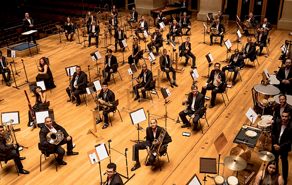 Imagem mostra sala de concerto com diversos músicos no palco olhando para a câmera