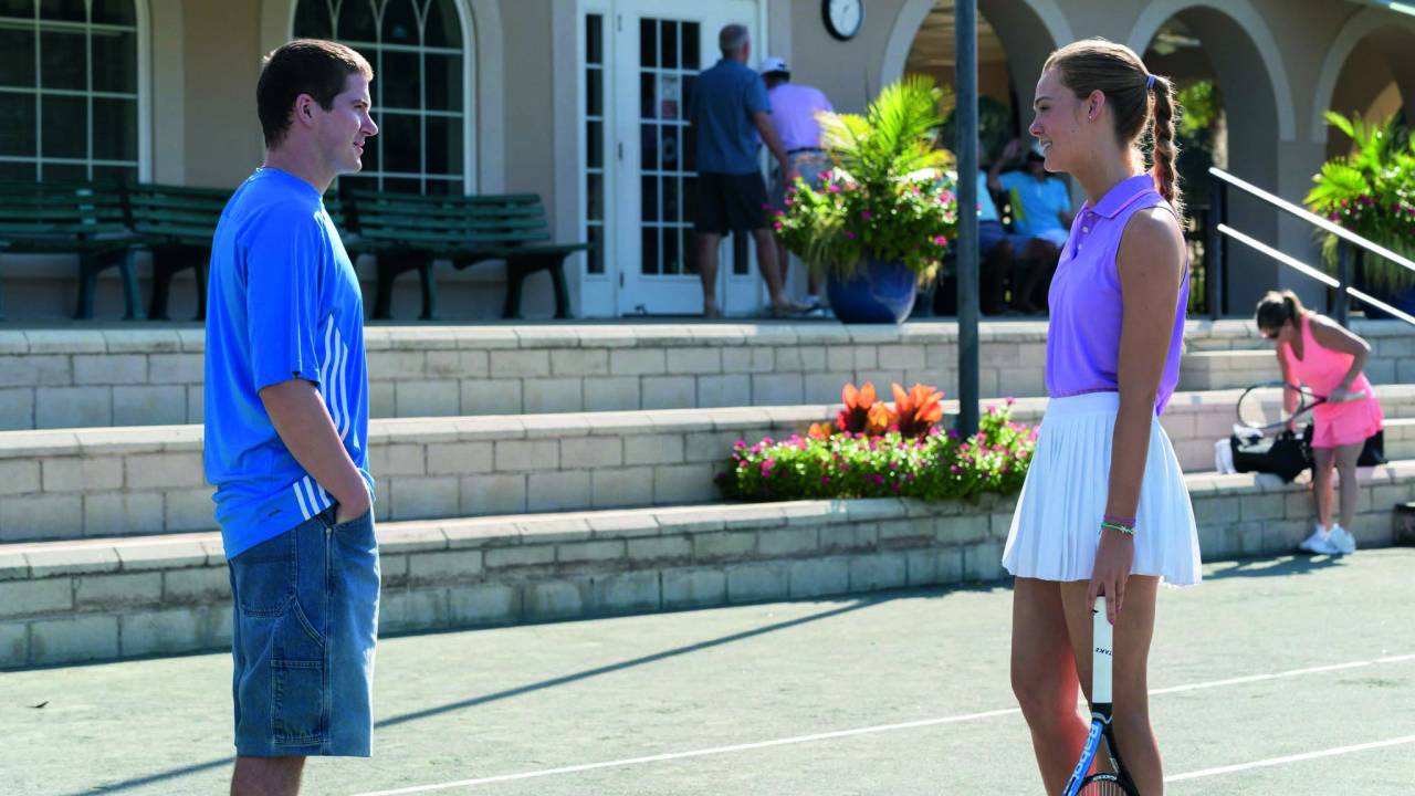Imagem mostra uma mulher com roupa de tenista e uma raquete na mão conversando com um homem em uma quadra de tênis
