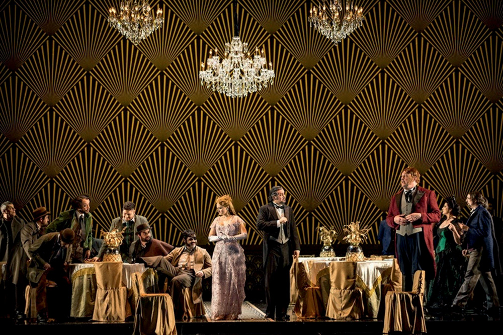 Imagem mostra diversos atores com roupas de gala em palco com cenário de jantar