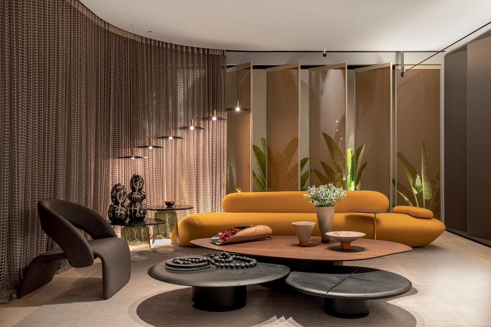 Sala de estar da CASACOR SP 2022. Em tons de marrom, bege, preto e laranja, tem um sofá arredondado laranja no centro e duas mesas de tampo redondo pretas logo a frente.