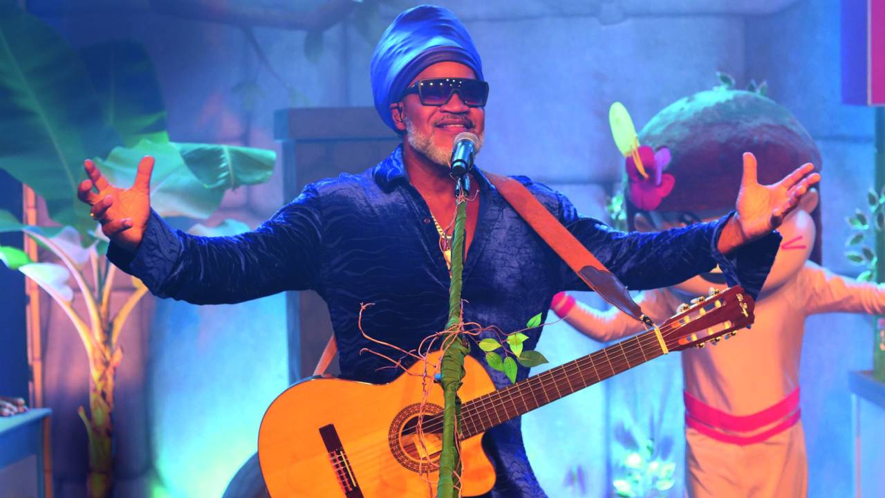 Imagem mostra homem em palco com os braços abertos, cantando. com um violão