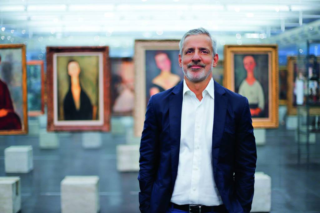 Imagem mostra homem de barba grisalha e camisa e terno em salão de museu. Ao fundo, diversas pinturas.