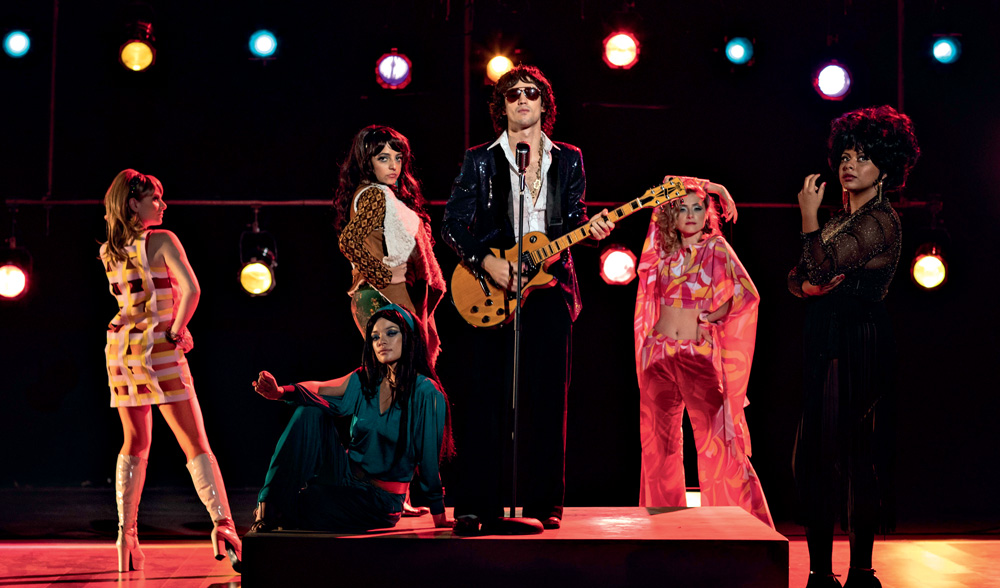 Imagem mostra homem em cima do palco segurando guitarra e cantando em microfone. Ao seu redor, diversas mulheres com roupas extravagantes
