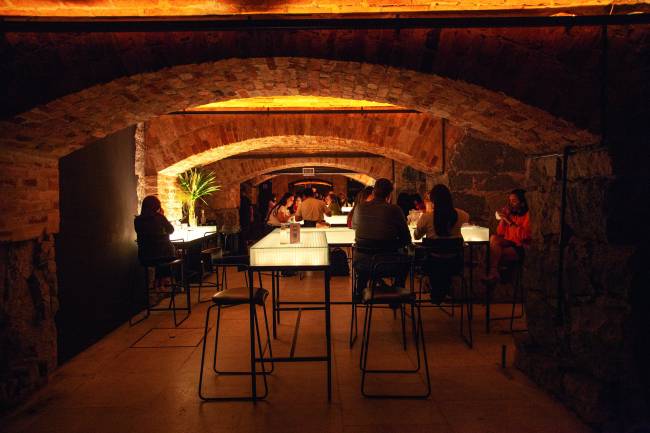 Imagem mostra interior de bar subterrâneo, com iluminação intimista e mesas brancas.