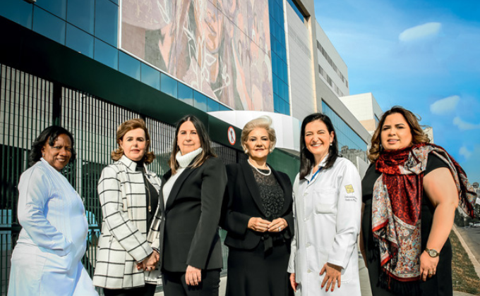 Imagem mostra seis mulheres enfileiradas, uma ao lado da outra, sorrindo, na calçada, em frente a hospital
