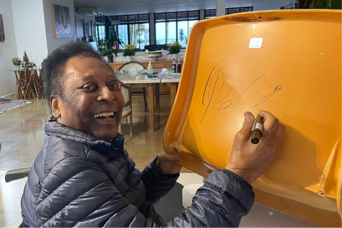 Pelé posa usando casaco e sorrindo, com mão sobre cadeira laranja do Pacaembu, apresentando sua assinatura