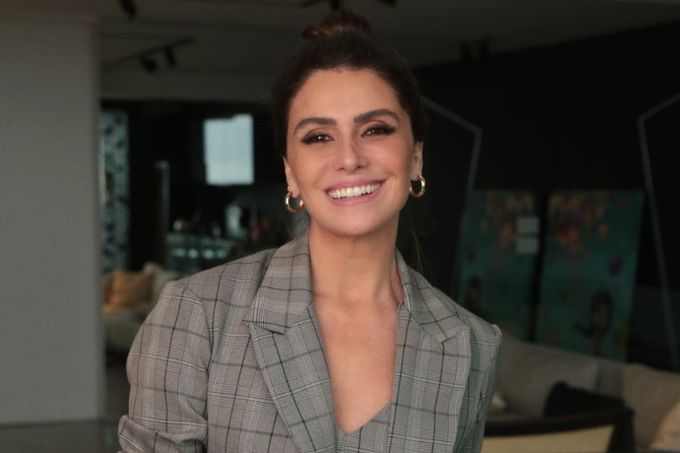 Giovanna Antonelli posa sorrindo e usando um blazer cinza xadrez, com sala de estar ao fundo