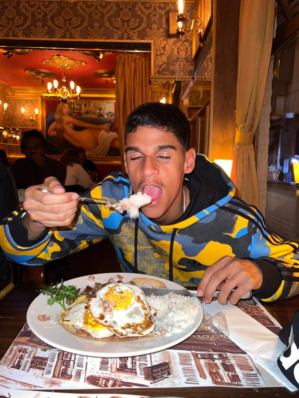 Luva de Pedreiro posa levando pedaço de comida à boca no restaurante Paris 6. À sua frente, prato com arroz, feijão e ovo frito
