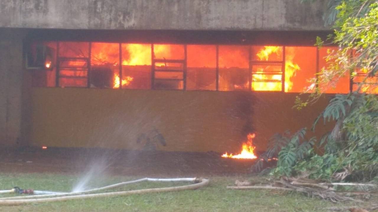 Foto mostra parte inferior de prédio com muitas chamas dentro. Do lado de fora, na grama, uma mangueira esguicha água para tentar controlar o fogo