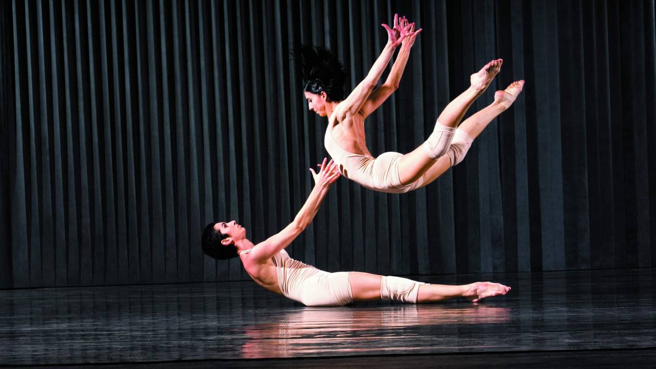 Imagem mostra dois bailarinos no palco. Um pula com os braços e pernas para trás, sobre o corpo do outro que está deitado