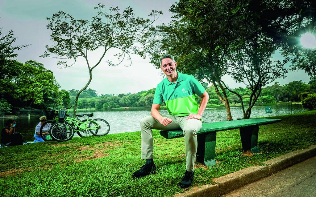 Imagem mostra homem de camiseta verde sentado em banco de madeira em parque. Ao fundo, um lago
