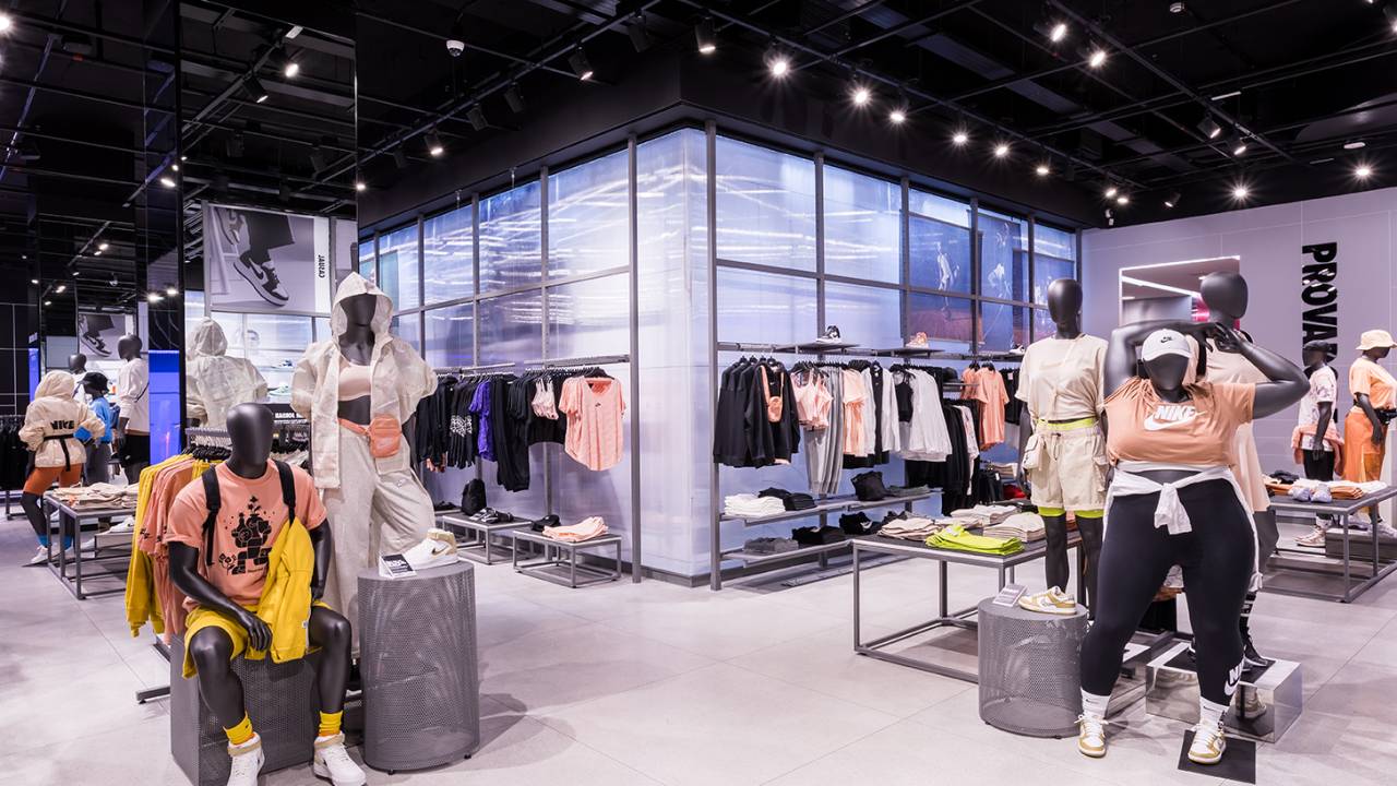 Foto exibe box de vidro no meio de loja moderna com chão cinza e manequins com roupas coloridas de atletismo.