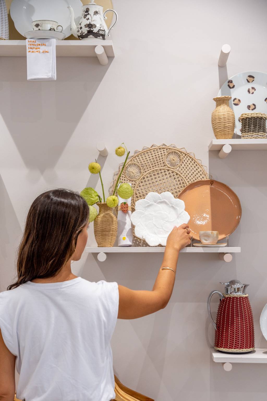 Mulher de costas aparece ajeitando prateleira com louças e vaso.