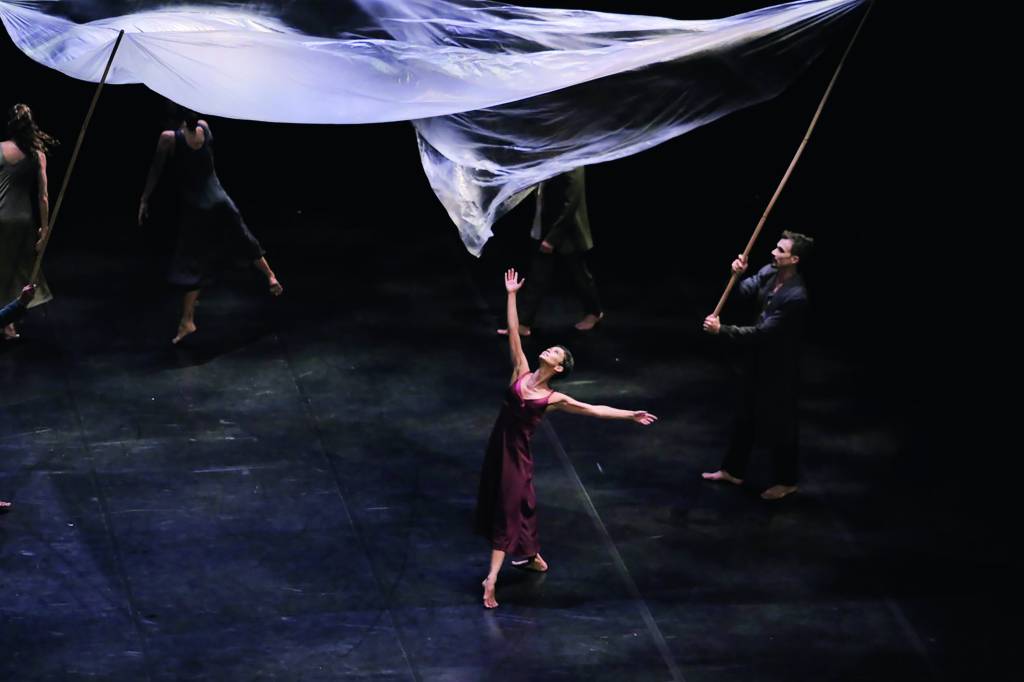 Dançarina no meio do palco, em movimento com os braços para cima. Usa vestido vinho reto e comprido e está rodeada por outros dançarinos que elevam panos brancos