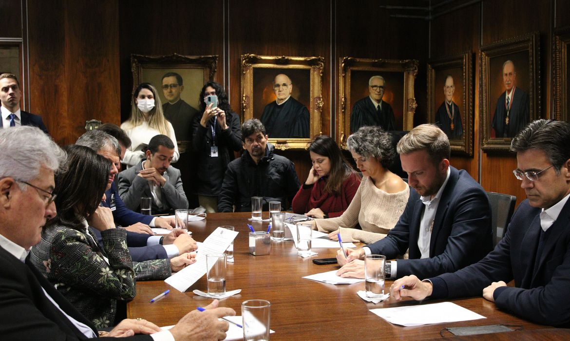 Foto exibe mesa com homens e mulheres olhando para documento.