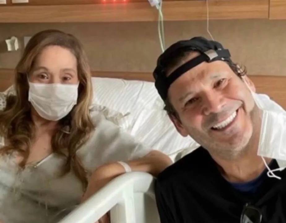 Imagem de Sonia Abrão sentada em cama de hospital usando máscara, ao lado de Elias Abrão, com máscara presa em apenas uma orelha, sorrindo para a câmera