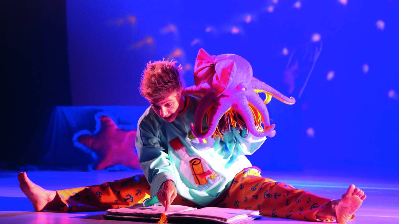 Imagem mostra dançarina abrindo espacate em palco, com figurino de criança