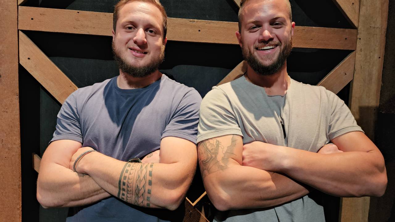 Dois homens posam de braços cruzados sorrindo. Ambos vestem camisetas escuras e estão em frente a estrutura de madeira.