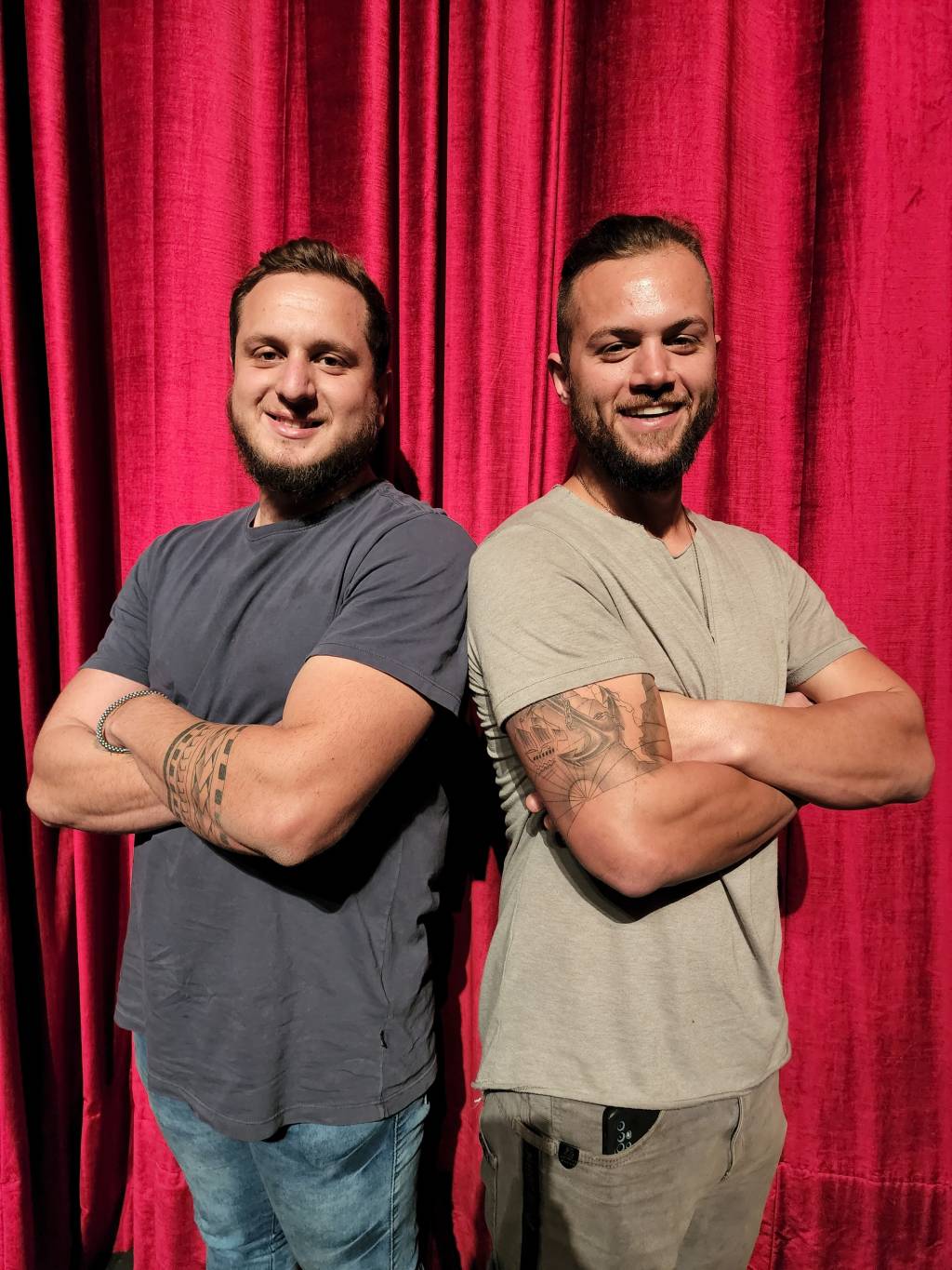 Dois homens posam de braços cruzados sorrindo. Ambos vestem camisetas escuras e estão em frente a cortina vermelha.