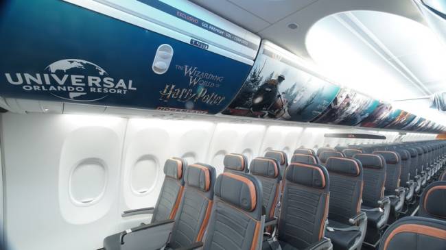 Foto de interior de avião, com quatro fileiras de cadeiras com três assentos. Na parte de cima, bagageiro com adesivo que mostra letreiro e cena de Harry Potter