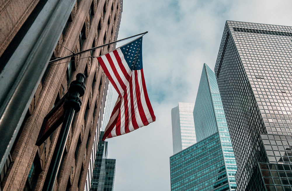 A low angle shot of the USA or United States of America flag on a flagpole near skyscrapers under a cloudy sky