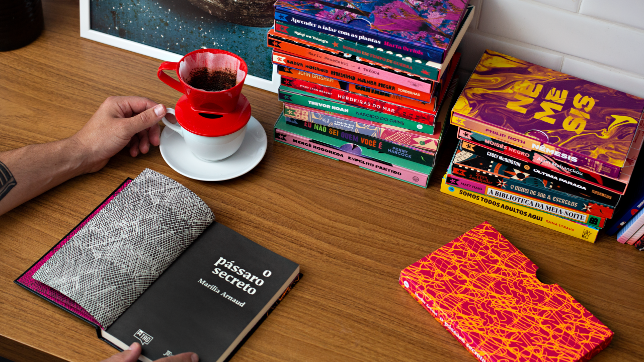 Foto exibe mesa de madeira com pilhas de livros coloridos, uma caneca de café branca e vermelha e duas mãos com um livro preto aberto.